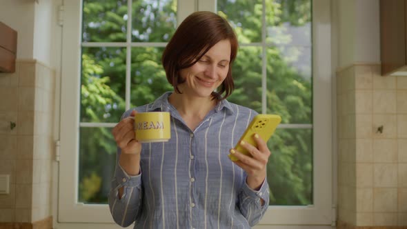 30s Woman Drinking Coffee of Yellow Cup with 'Dream' Sing Using Mobile Phone Standing in the Kitchen alt