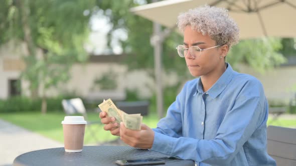 Young African Woman with Coffee Counting Dollars in Outdoor Cafe alt