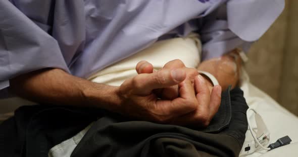 A middle aged male medical patient sits in a hospital bed rubbing his hands together nervously while alt