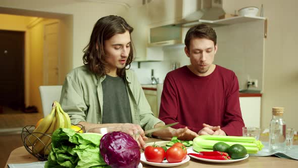 A Young Man is Cutting Cucumbers for a Salad and Talking to His Friend