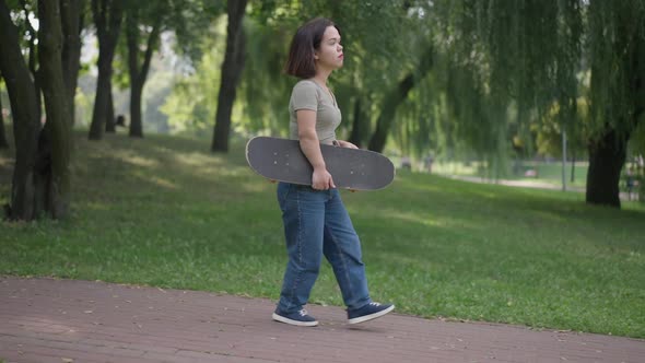 Side View Young Caucasian Beautiful Woman with Dwarfism Strolling with Skateboard in Park alt