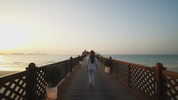 Woman Walking on the Wooden Pier During Sunset with Dubai Skyline in the Background alt