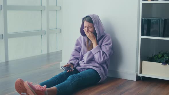 Sad Young Woman Sits on the Floor Looking at Cellphone Screen and Begin Cry alt