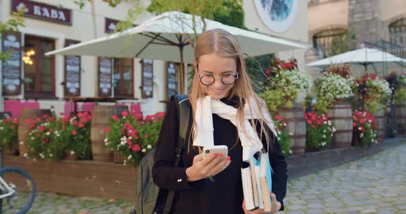 Young Woman in Glasses which Using Her Phone while Standing Near Summer Outdoors Cafe alt