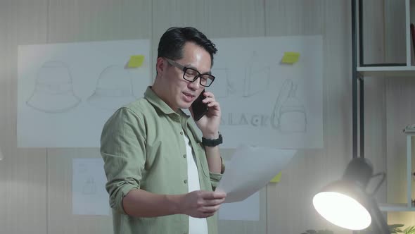 Asian Man Bag Designer Talking On Smartphone While Looking At The Layout Bond In Hand And Walking alt