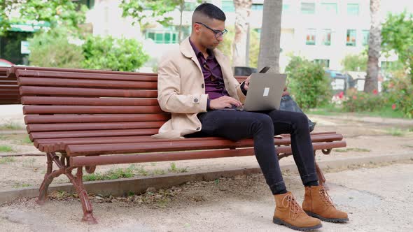 Young businessman in trendy outfit speaking on phone on bench alt