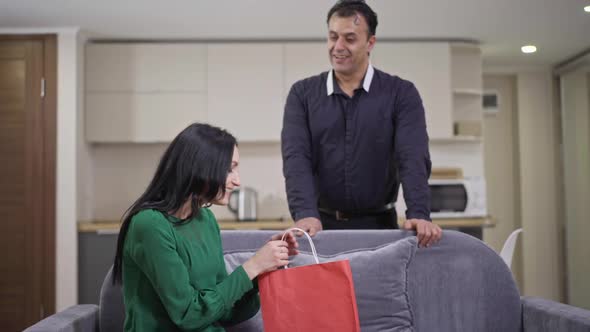 Smiling Wealthy Middle Eastern Man Giving Gift to Surprised Excited Caucasian Woman Sitting on Couch alt