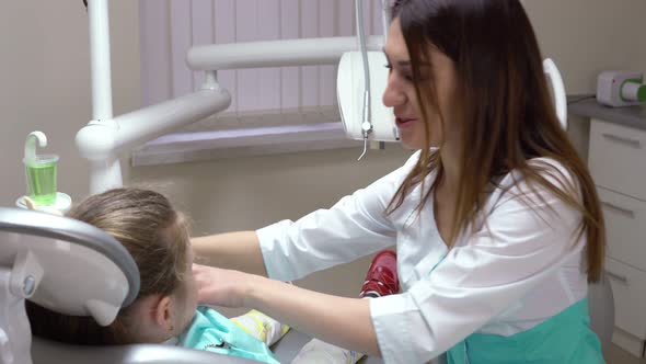 Friendly Female Dentist Putting a Napkin on the Cute Little Girl Taking on Gloves and Protective alt