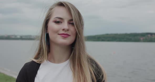 Cheerful Girl Corrects Her Blowing Hair and Smiles at Camera at Seaside Walk alt
