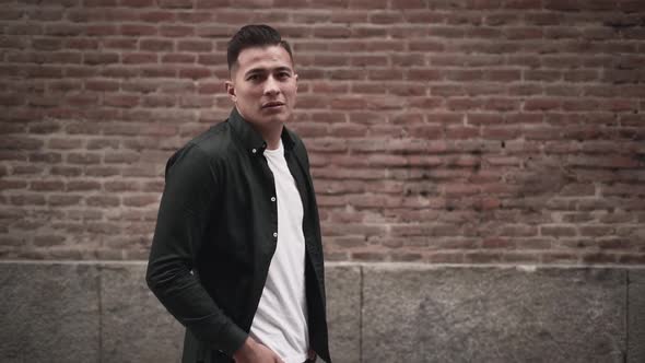 Hispanic Young Man Looking at the Camera and Posing with Hands in Pockets in Front of a Brick Wall alt
