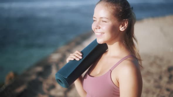 Delighted Nice Girl with Ponytail Holds Yoga Mat on Beach alt