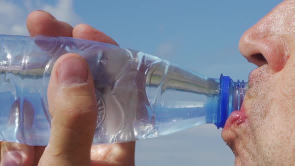 A Man Drinks Water From a Transparent Plastic Bottle Against a Blue Sky alt