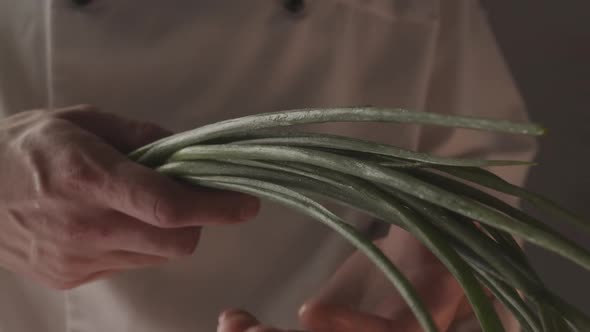 Chef in Uniform Works in the Kitchen and Holds Green Onions in His Hand alt