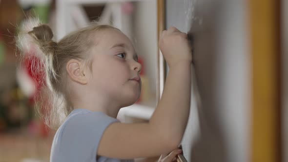 Side View of Genius Little Girl Writing with Chalk on Blackboard in Classroom alt
