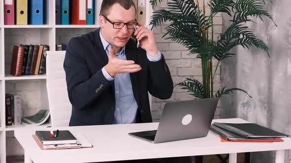 Disgruntled Frustrated Businessman in Glasses and Jacket Talking on Mobile Phone Sitting in Office