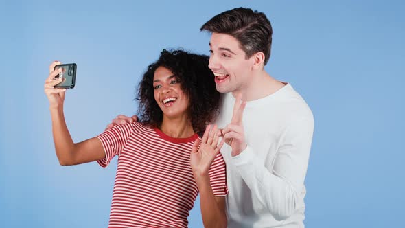 Young Interracial Couple Making Selfie with Smartphone on Blue Studio Background alt