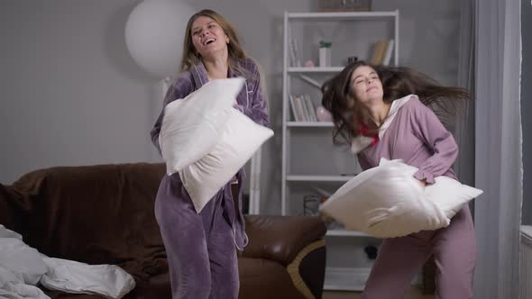 Two Joyful Caucasian Young Women Dancing with Pillows at Pajama Party Indoors alt