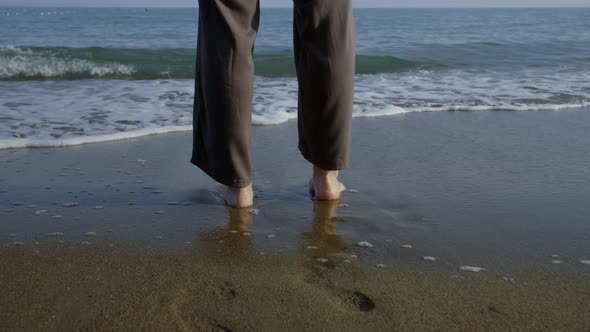 Woman Is Standing in Water of Sea on Sandy Beach, Waves Are Washing Her Feet alt