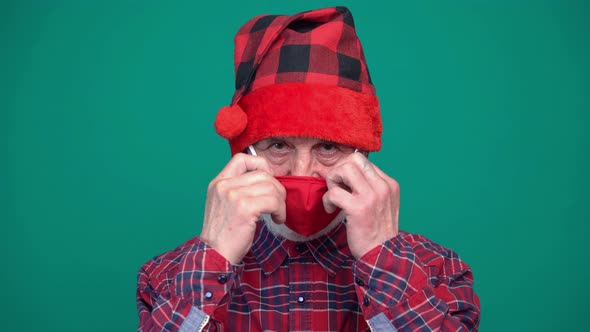 Portrait of Mature Man in Santa Hat Wears a Red Medical Mask Looking at Camera