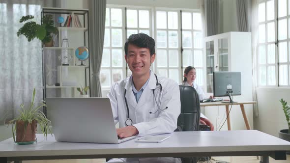 Young Asian Man Doctor Is Using Laptop Computer, He Turns And Smiles To Camera In Workplace alt
