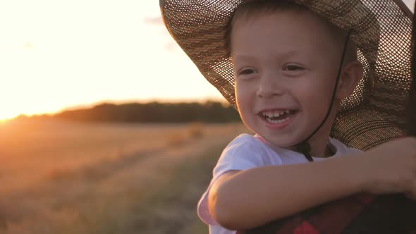 Happy Little Toddler Boy in Hat Having Fun Sitting in Parents Arms in Field in Summer at Sunset alt