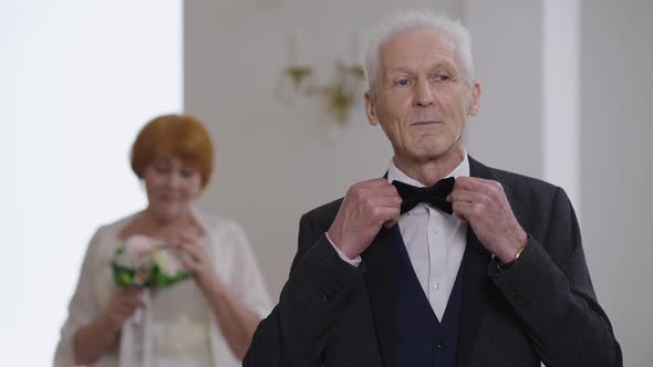 Confident Caucasian Greyhaired Groom Adjusting Bow Tie Looking at Camera Smiling alt