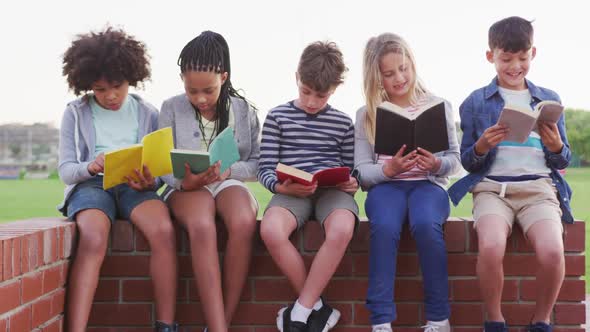 Group of kids reading books while sitting on a brick wall alt