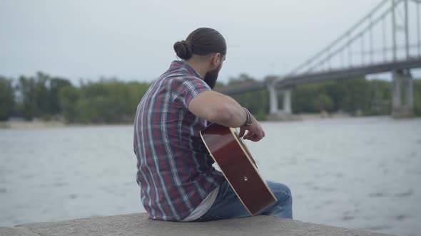 Back View of Absorbed Caucasian Guitarist Playing on River Bank with Blurred Bridge at the alt