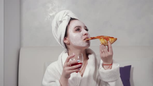 Girl in a Towel on Her Head and in a White Robe Drinks Red Wine and Eats Pizza