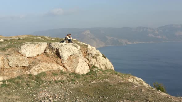 People Sit and Eat on Steep Rocky Cliff Edge Against Coastline alt