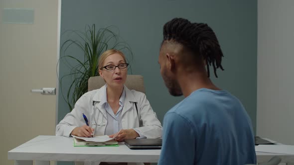 Female Doctor Writing Out Medical Prescription to Male Patient Indoors alt