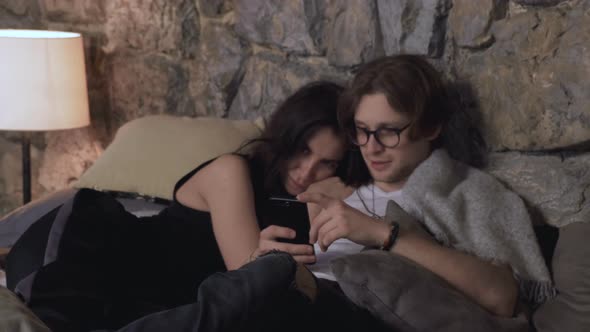 Camera In Motion A Young Couple Are Lying In Bed Chatting Looking At Photos On Their Smartphone  alt