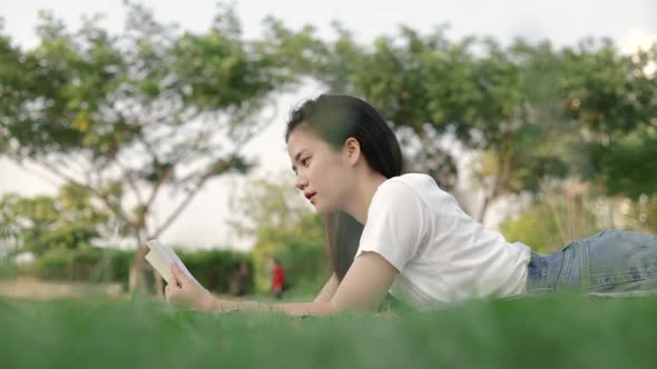 A beautiful Asian woman relaxed reading a book while lying on the floor grass outdoors. alt