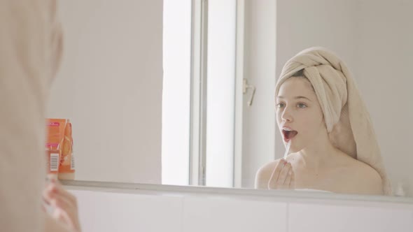 Teenage girl applying lip balm and lip gloss in front of the bathroom mirror alt