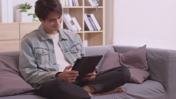 sian man sits on sofa at home, texting and talking with buddy, holding smartphone,or checking mobile alt