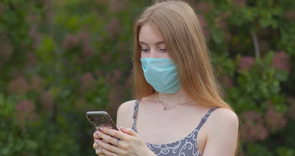 A young girl in a protective medical mask looks at the phone and sends messages