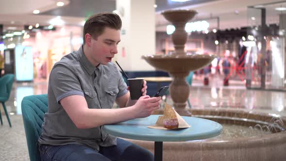 A Young Man in a Cafe with a Phone in His Hands Against the Backdrop of a Fountain alt