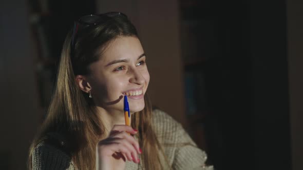 Happy Girl with Pen in Hand Cute Smiling at Camera at the Desk in Dim Library alt