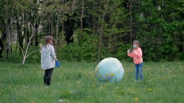 Two Girls in Medical Masks Play with a Large Inflatable Ball in the Park in the Spring. The Concept
