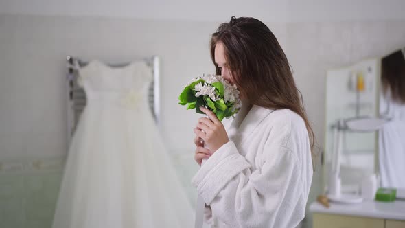Side View of Young Caucasian Bride Smelling Bridal Bouquet Admiring Wedding Dress Standing in alt