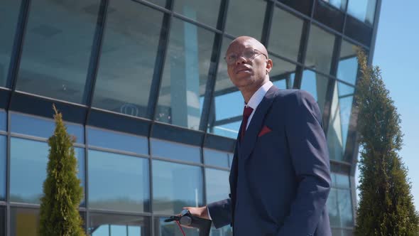 A Young Business Man in a Classic Blue Business Suit Stands with an Electric Scooter on the Street alt