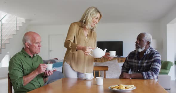 Two diverse senior couples sitting by a table drinking tea together at home alt