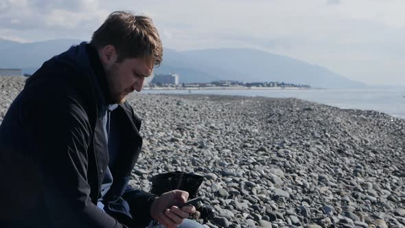 Man Traveler Sits on a Stone Beach of the Sea and Looks Through a Smartphone