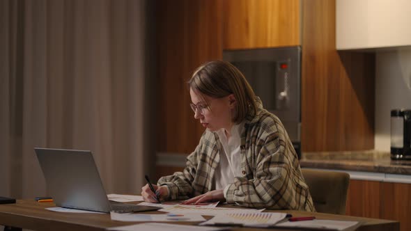 Woman with Glasses Working Remotely in a Home Office at a Desk with a Laptop and Notes Data on a alt