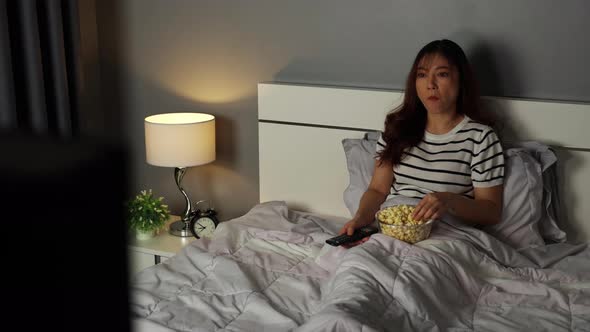 young woman watching TV and eating popcorn on a bed at night
