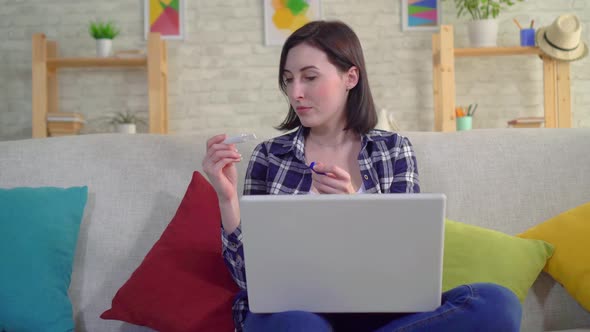 Young Attractive Smiling Woman with Pregnancy Test Sitting on Sofa with Laptop alt
