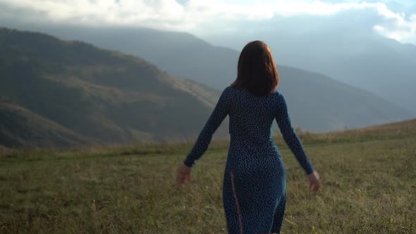 A Young Woman Walks in a Blue Dress and Raises Her Hands Up, Looking at the Mountains. alt