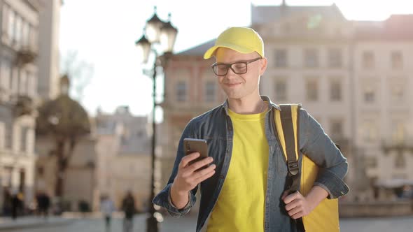 Portrait of a Happy Young Delivery Man Who is Going and Texting on the Smartphone alt