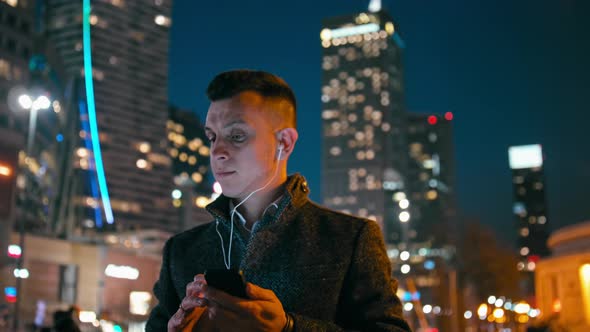 Man with Earphones Is Listening To Music on Phone in City at Night with Lights alt