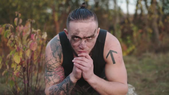 Brutal Athlete with Soot on Face and Tattooes Leans on Knees and Looks at Camera alt
